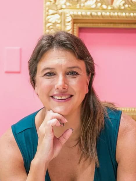 Jess Laverde smiling, chin on her hand, in front of a pink wall and gold frame